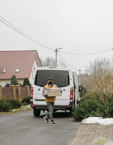 A delivery person wearing a mask carries a box from a van in a residential area.