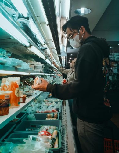A young man wearing a mask shops in a supermarket refrigerated aisle, selecting groceries.